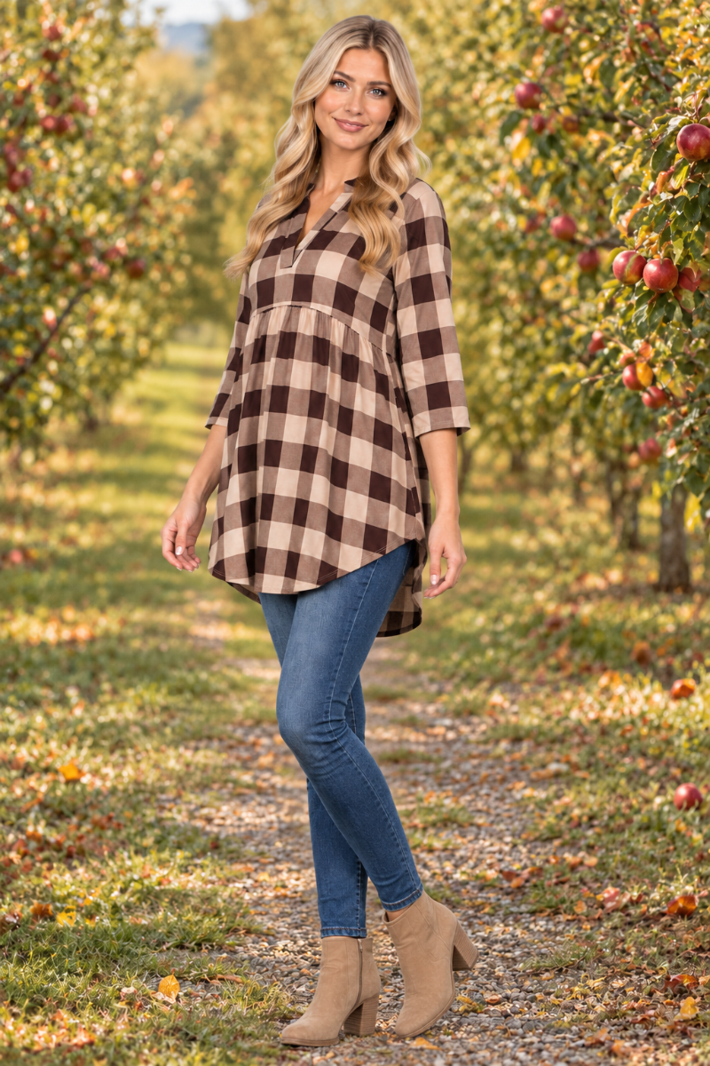 Full-body half-side lifestyle view of a blonde model wearing the beige-and-brown plaid tunic, jeans, and tan boots in an apple orchard