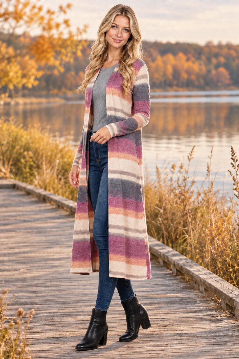 Full-body half-side lifestyle view of a blonde model wearing the striped cardigan, gray top, jeans, and black boots beside an autumn lake