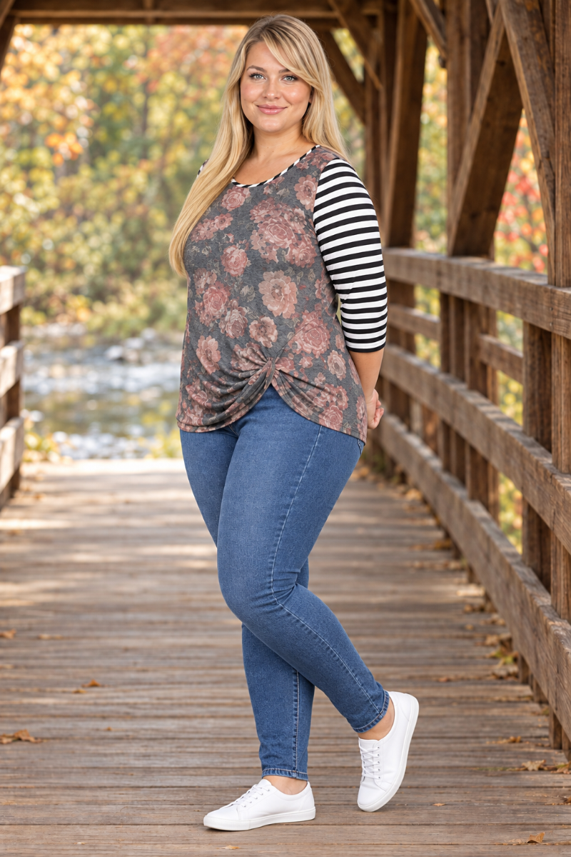 Full-body half-side lifestyle view of blonde model in charcoal floral top with striped sleeves and blue jeans on a covered bridge