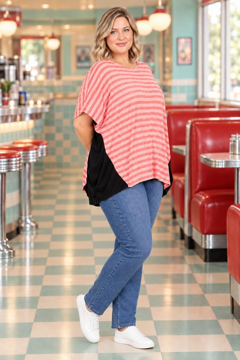 Full-body half-side lifestyle view of blonde model in coral-striped dolman top with black side panels and blue jeans in a retro diner