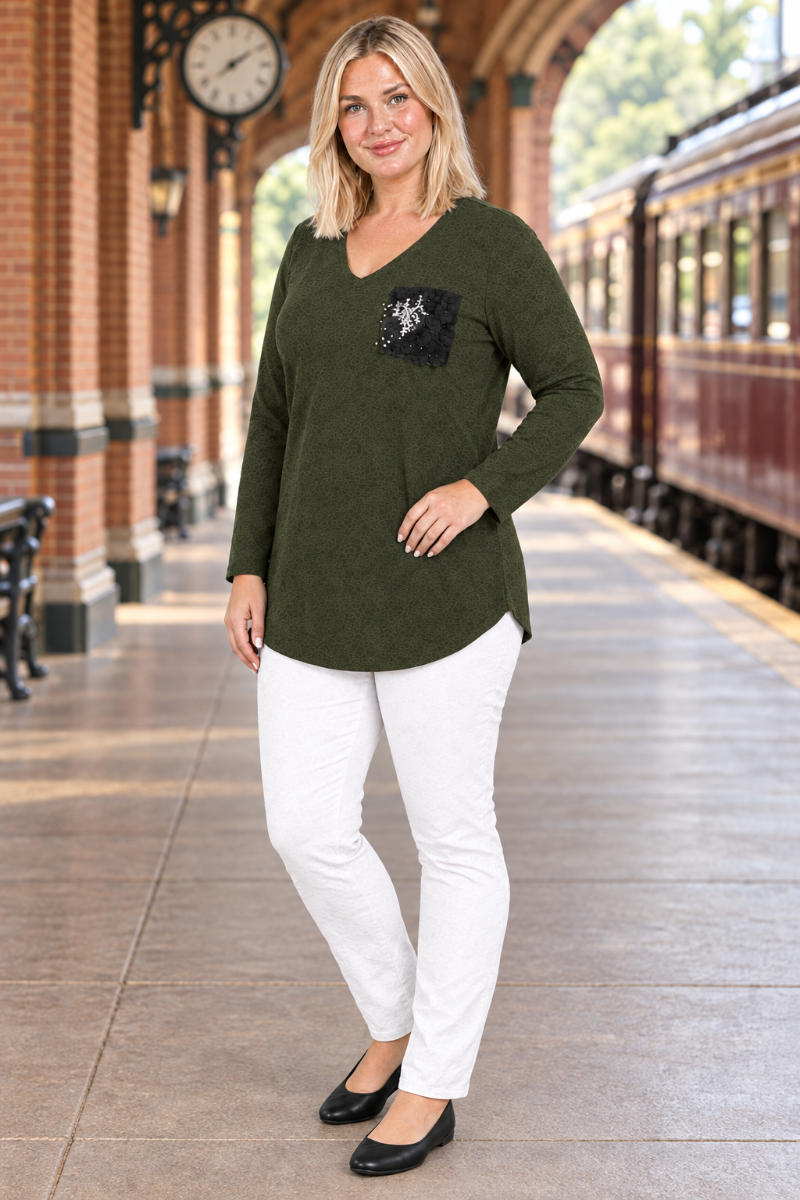 Full-body half-side view of blonde model in dark olive tunic with black embellished pocket, white pants, and black flats at a historic train station
