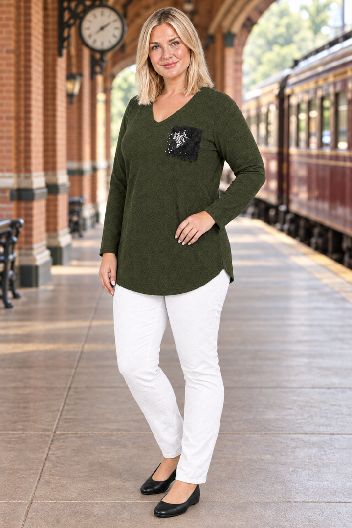 Full-body half-side view of blonde model in dark olive tunic with black embellished pocket, white pants, and black flats at a historic train station