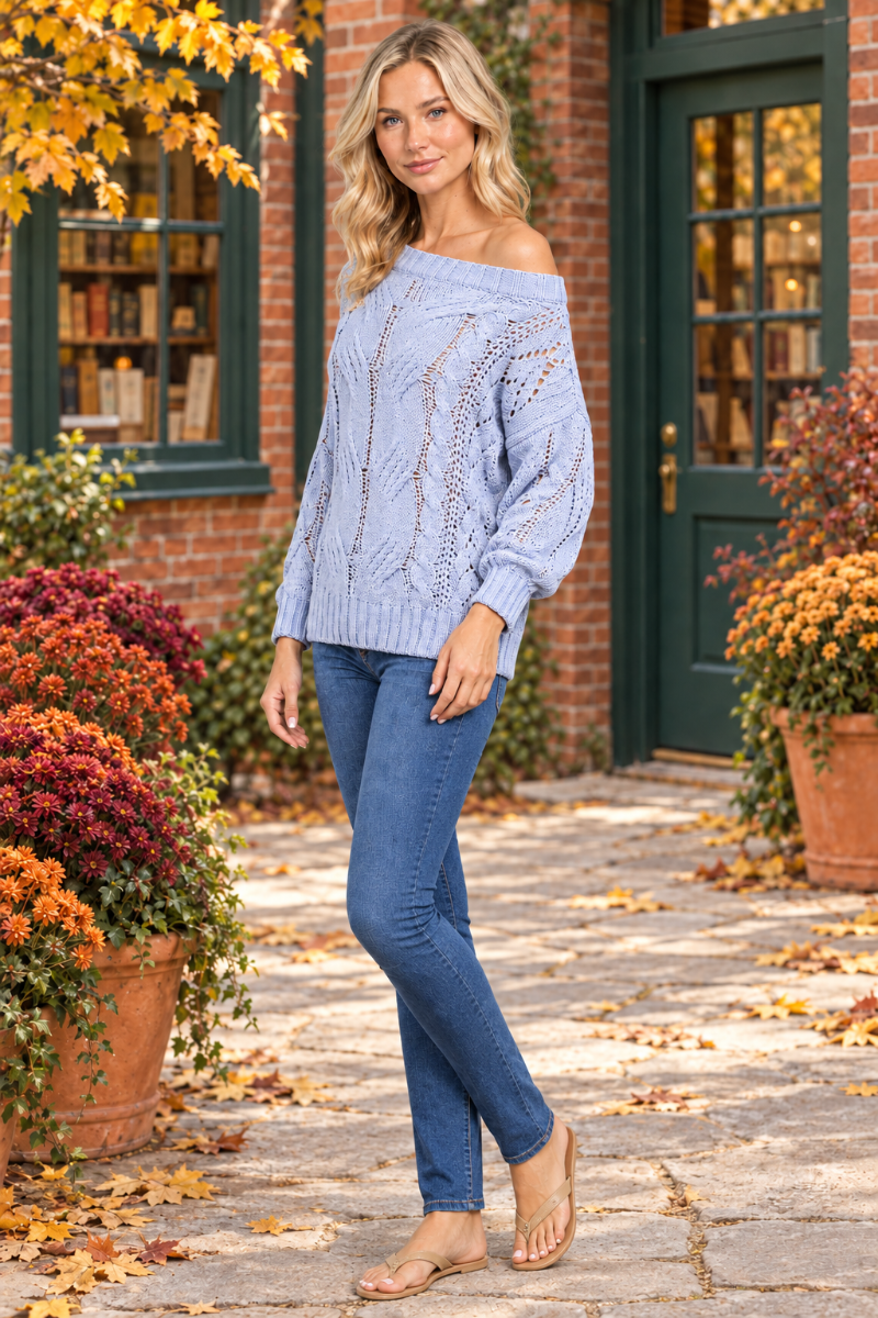 Full-body half-side view of the blonde model wearing the pale-blue open-knit sweater and jeans in an autumn courtyard