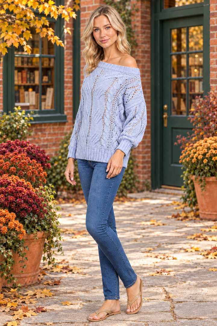 Full-body half-side view of the blonde model wearing the pale-blue open-knit sweater and jeans in an autumn courtyard