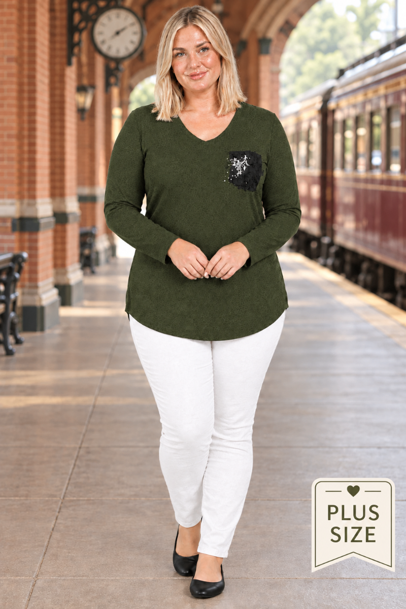 Full-body lifestyle front view of blonde model in dark olive tunic with black embellished pocket, white pants, and black flats at a historic train station