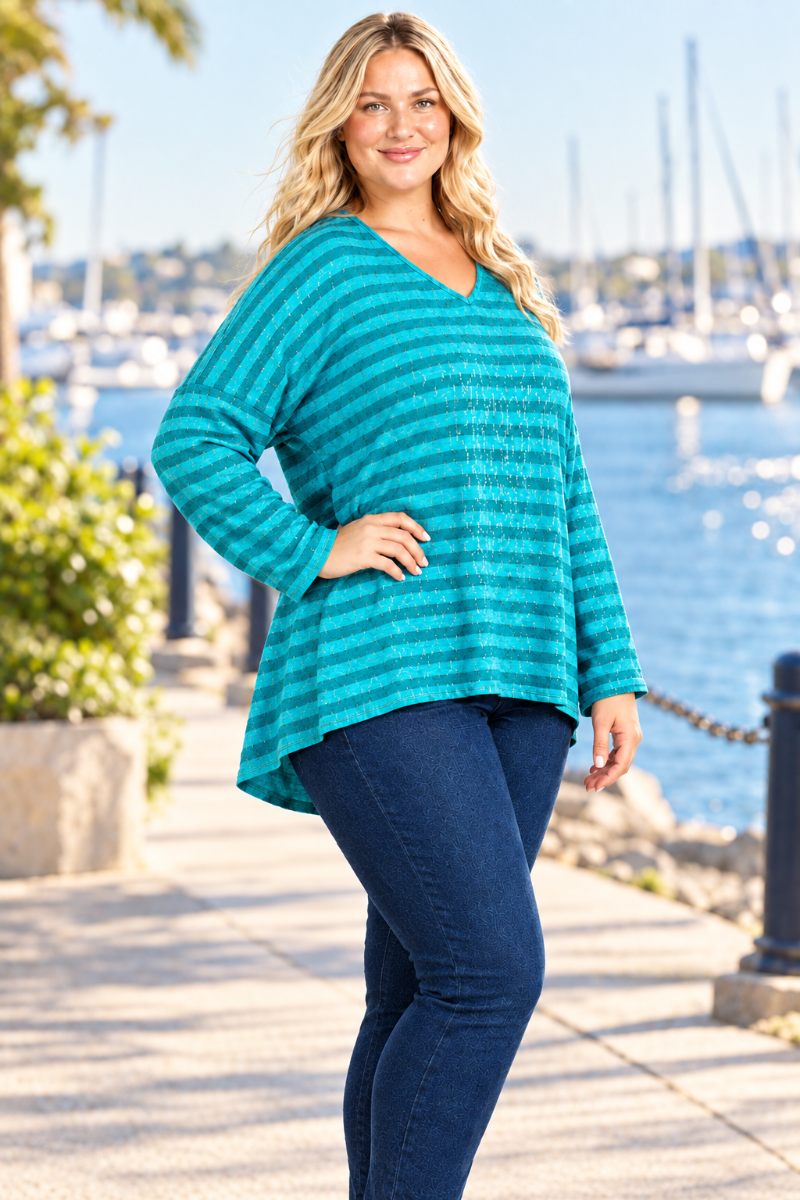 Half-side lifestyle view of blonde model in a loose teal striped top and dark jeans, harbor background