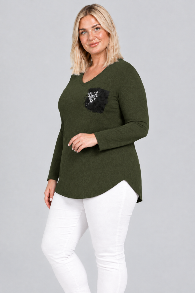 Studio side view of blonde model in dark olive V-neck tunic with black embellished pocket and white pants