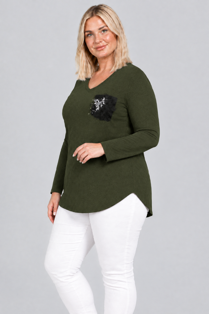 Studio side view of blonde model in dark olive V-neck tunic with black embellished pocket and white pants